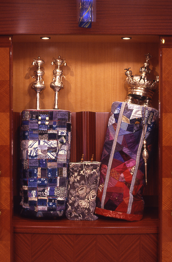 "Hidden and Revealed", collaboration with Dorothy Ross, fabric and beadwork construction. Commissioned by Women for Reform Judaism for the Golomb Chapel at URJ (New York NY). Just as the tightly structured wedding kimono has, surprisingly, a crimson silk lining, so it is a joy to discover delights within the traditional and sometime formulaic words of Torah.This mantle wraps the scroll in an assembly of square parcels. In Buddhist and Shinto belief squares connote completeness and balance. Silk-screened on some squares are texts referring to God's truths being both hidden and revealed. The complete texts are scrolled into the breastplate, like private treasures within a locket. This piece is fastened with rows of five pearls, five being a number associated in Judaism with the senses, creating, and protective hamza. The obi [sash] area holds a beaded yad [pointer used while reading the scroll].