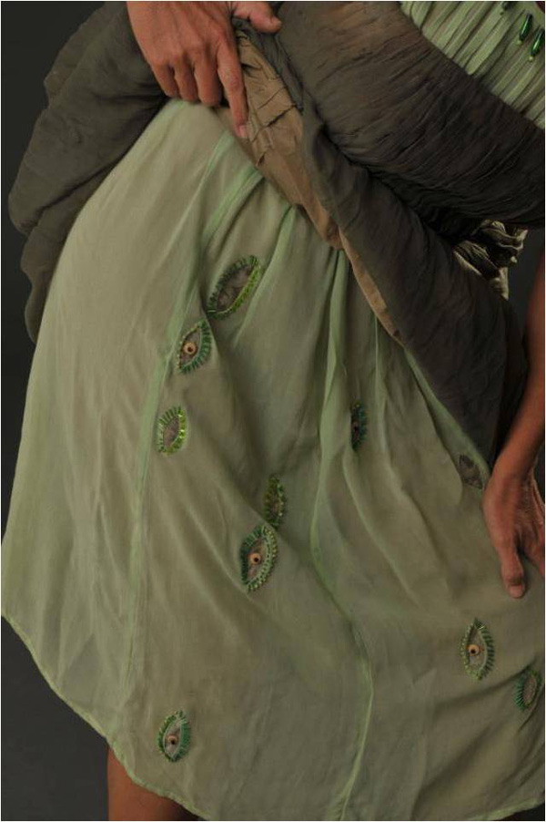 ARTEMISAL/LASTIMARÉ (ARTEMISIAL/WILL HURT) is a dress dedicated to the virgin goddess Artemisia/Diana, inflexible Lady of wild beasts, jealous guardian of her own freedom, the one who isn’t owned. Careful! She is potentially dangerous to her offenders.
An antique iridescent satin ball gown, overlapping a girl’s dress, in which skirt vertical eyes have been carved in tapestry brocade and embroided with small glass beads. Both dresses are green, although in different shades; both have the same ornamental treatment in their chest, but one is machine-sewn and the other was hand sewn and worn by my mother and her two elder sisters, stained with anticipation and transpiration. The palimpsest of both dresses (with a silver crescent moon tattooed on its right shoulder) makes an offering to the virgin goddess Artemisia, patron of young girls, savage beauty, and freedom in wilderness.
Materials: Two vintage silk dresses; used brocade tapestry; colored glass beads; silver thread. Hand sewn.
Photo: Maurizio Donelli.