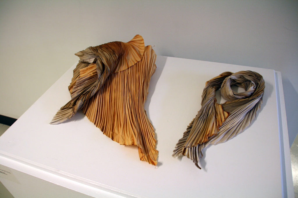 Left: Alight, 2016. 40 H X 18 W X 9 D cm, cotton fabric sculpture, Technique: Arashi Shibori dyed with rust & tea, displayed lying on wooden or white surface. Long deeply ridged fabric twisted slightly & coiled with ends overlapping.
Right: Winged, 2016. 52 H X 28 W X 7 D cm, cotton fabric sculpture, Technique: Arashi Shibori dyed with rust & tea, displayed lying on wooden or white surface. Long deeply ridged fabric folded in two with top end slightly shorter & twisted inward.